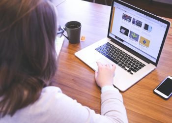 Person browsing the web on a MacBook at a wooden desk with a smartphone.